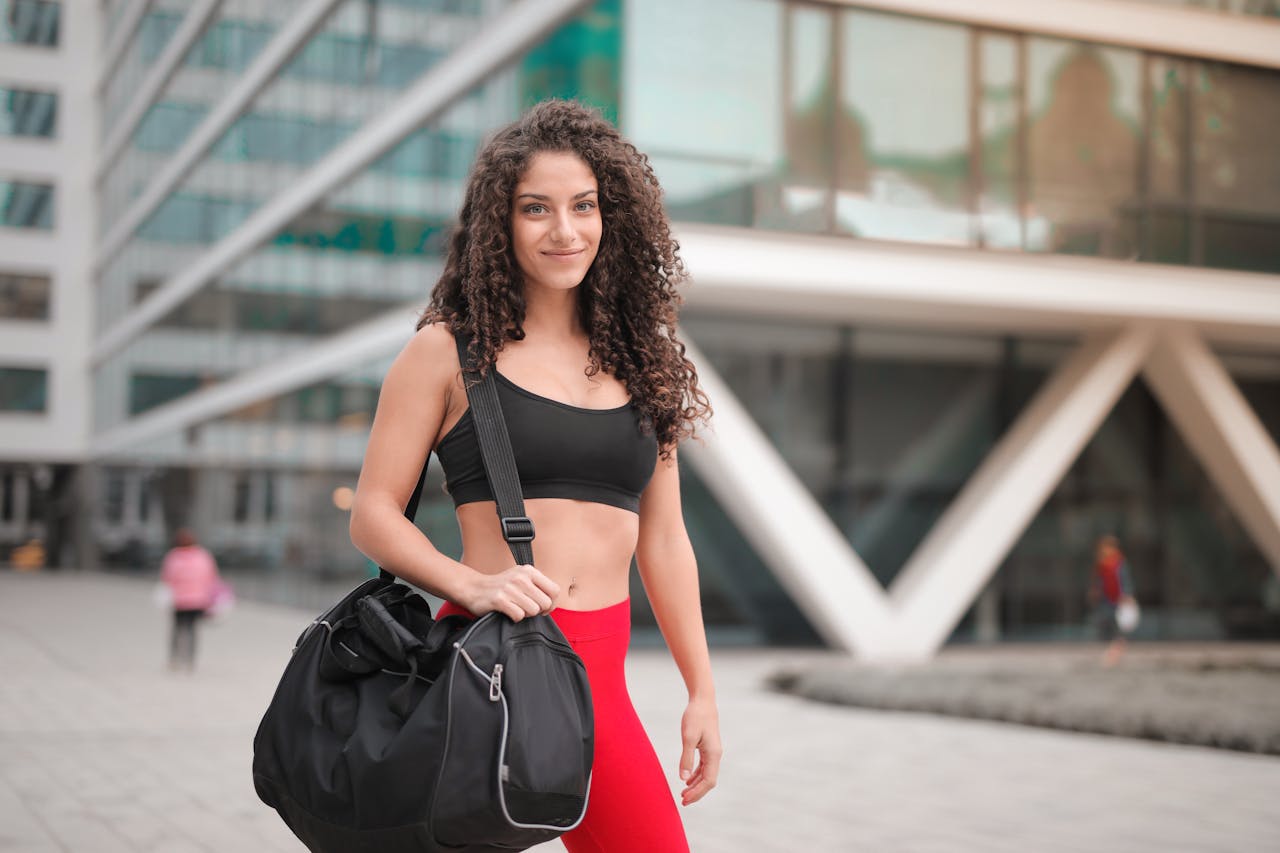 Young woman in activewear smiling with a gym bag outdoors in urban setting.